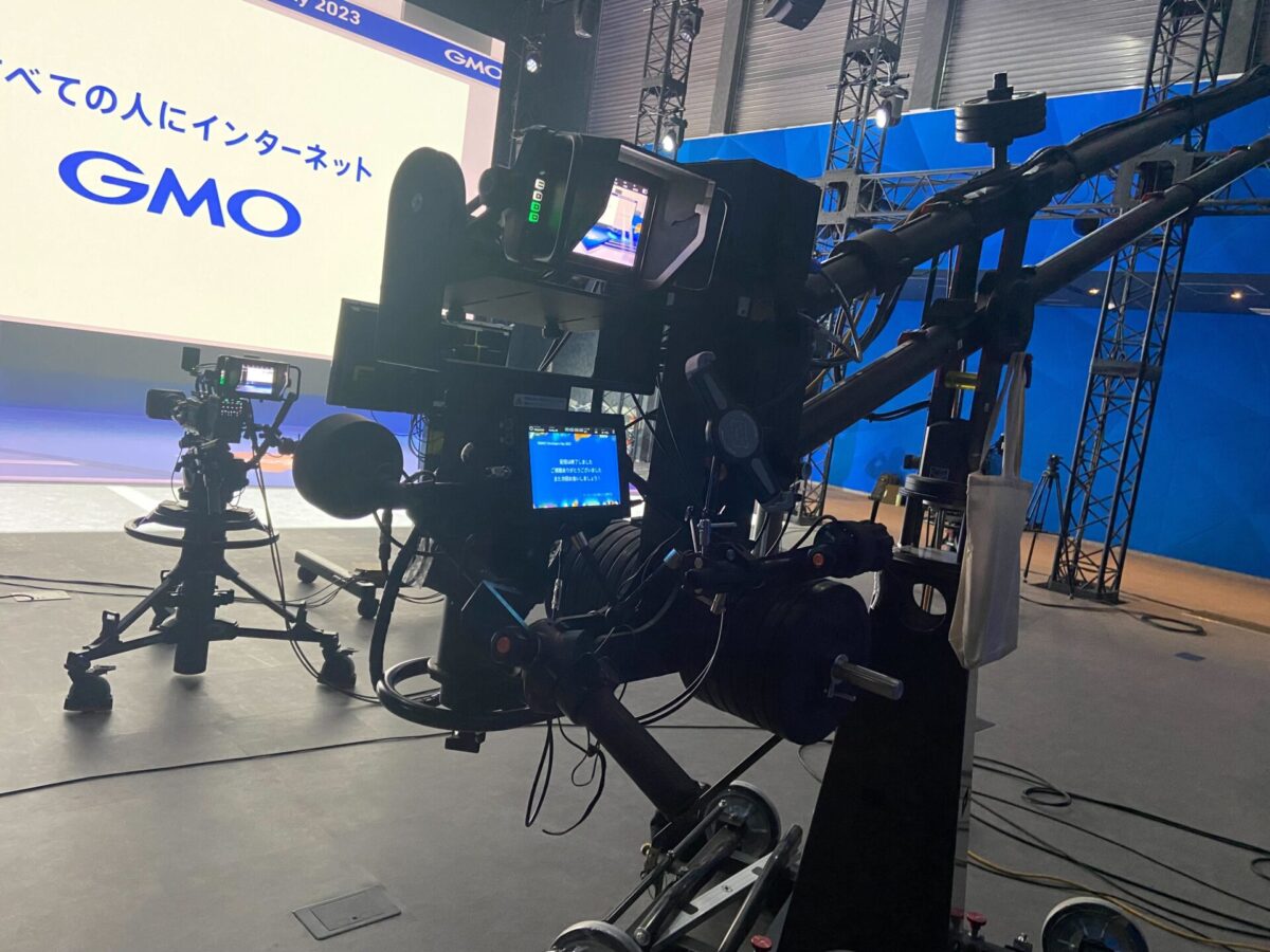 GMOグローバルスタジオに設置された大型カメラジブ（クレーン）システム。手前にはカメラのパン・チルトやズームを操作するモニターとコントローラーが配置され、奥にはもう一台のカメラと「GMO」のロゴが表示された背景スクリーンが見える。プロフェッショナルな配信・収録環境の様子。